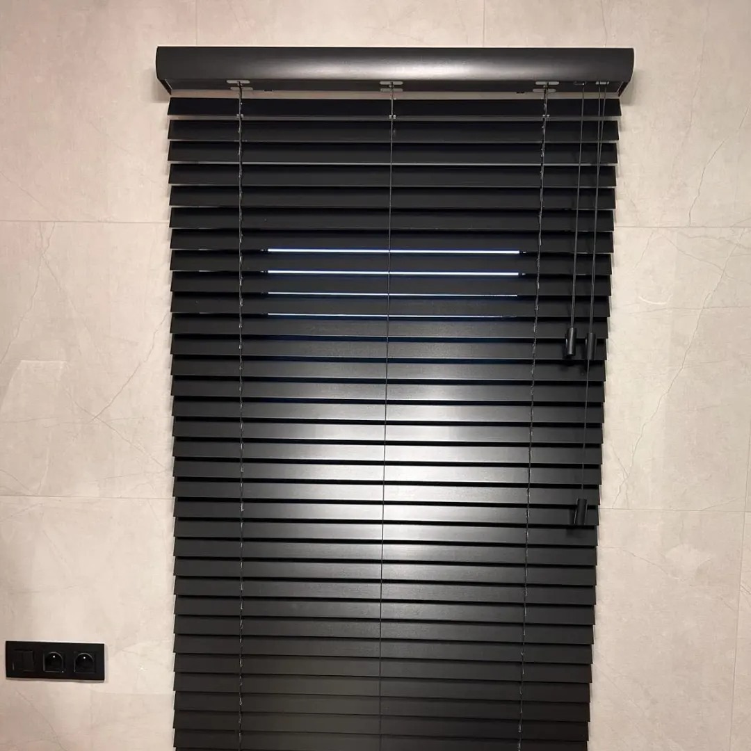 Czarne żaluzje poziome z listwami, częściowo odsłonięte, zamontowane na jasnej ścianie z marmurowym wzorem, obok włącznika światła.