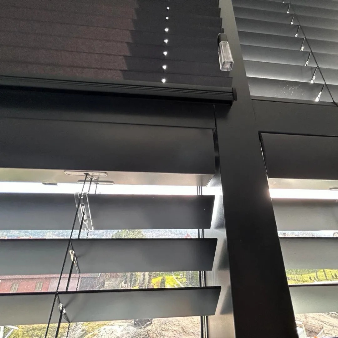 Ciemne żaluzje poziome, częściowo otwarte, z widokiem na zewnątrz. Detal mechanizmu i sznurków. Ujęcie z dołu, podkreślające strukturę i kolor.