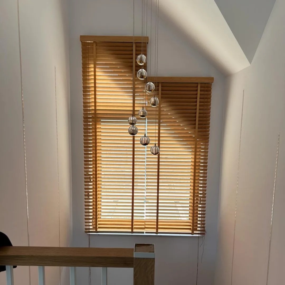 Drewniane żaluzje poziome w oknie o nieregularnym kształcie na poddaszu, z dekoracyjną lampą wiszącą z kulami. Widok z klatki schodowej.