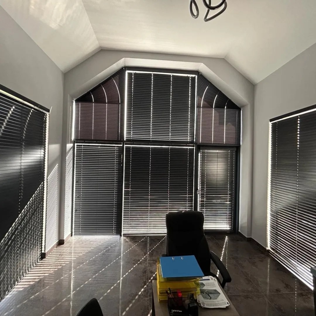 Eleganckie, czarne żaluzje poziome w nowoczesnym biurze z oknami o nietypowym kształcie. Światło słoneczne tworzy wzory na podłodze. Widok na biurko.