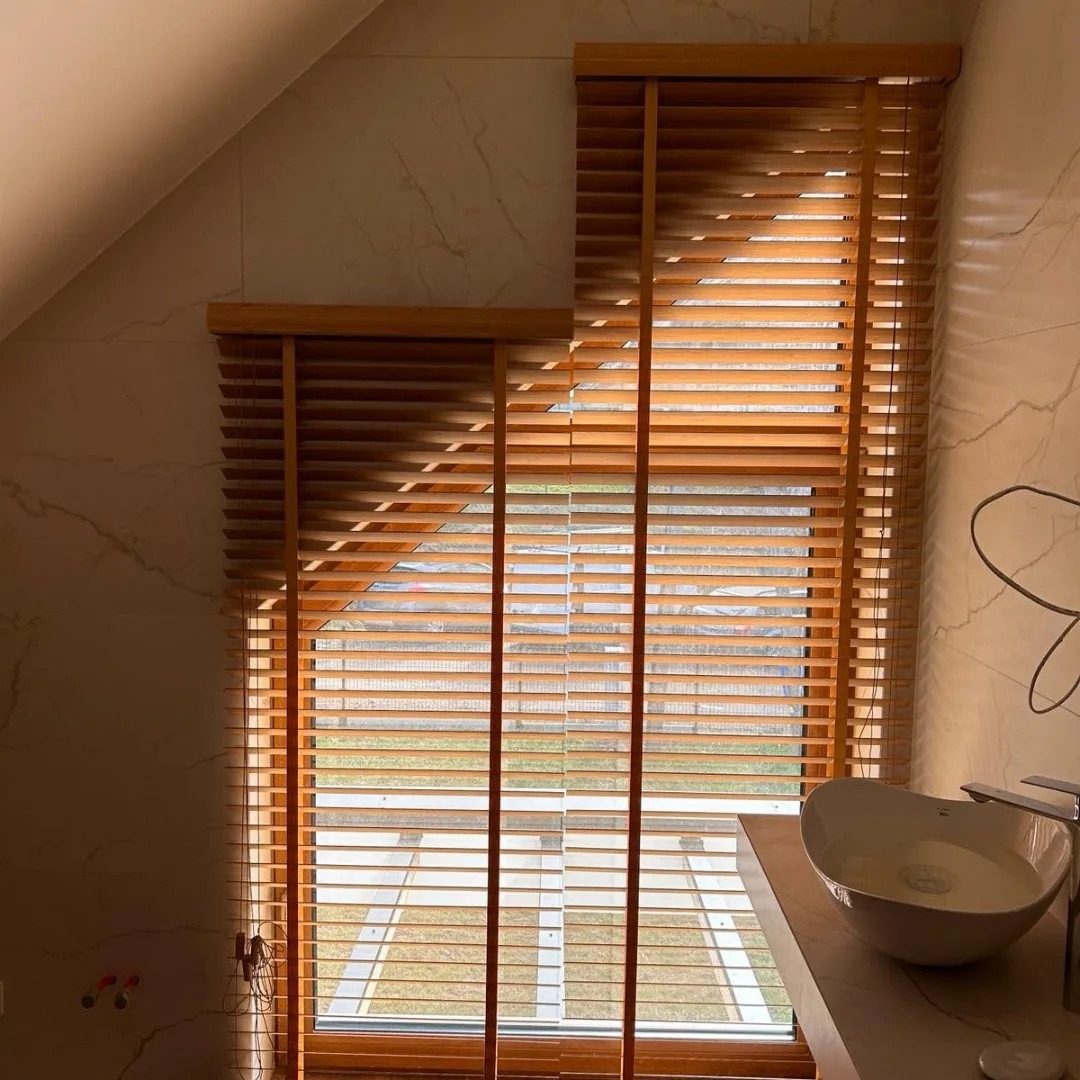 Drewniane żaluzje poziome w łazience z marmurowymi płytkami, widok na zewnątrz przez okno, ukośny sufit, ciepłe oświetlenie.