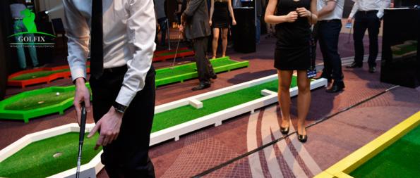 Mobilny tor do mini golfa z graczami w tle, widoczny fragment kija golfowego, kolorowe przeszkody i sztuczna trawa. Atrakcja na event firmowy.