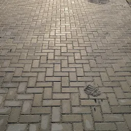 Długa, nowo ułożona ścieżka z szarej kostki brukowej ułożonej we wzór jodełki, z widocznymi dwoma studzienkami kanalizacyjnymi i śladem buta w piasku na powierzchni.