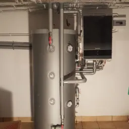 Szare zbiorniki akumulacyjne ciepła z podłączonym kotłem gazowym Buderus, widoczne rury i zawory, instalacja w piwnicy wykończonej płytkami.