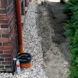 Drenaż opaskowy budynku w trakcie realizacji: rura drenarska, żwir, wykop. Widoczny budynek z cegły i zieleń. W tle drzewa.