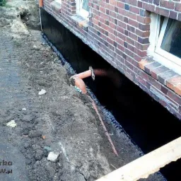 Odkopane fundamenty domu z cegły, zabezpieczone czarną izolacją. Widoczna rura drenażowa i częściowo zasypana ziemia. Prace ziemne w toku.