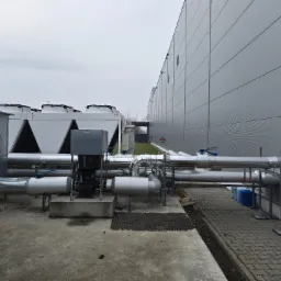 Instalacja przemysłowa z rurami i urządzeniami wentylacyjnymi na dachu budynku. Widoczne elementy chłodnicze i wentylatory. Szara kolorystyka i pochmurne niebo.