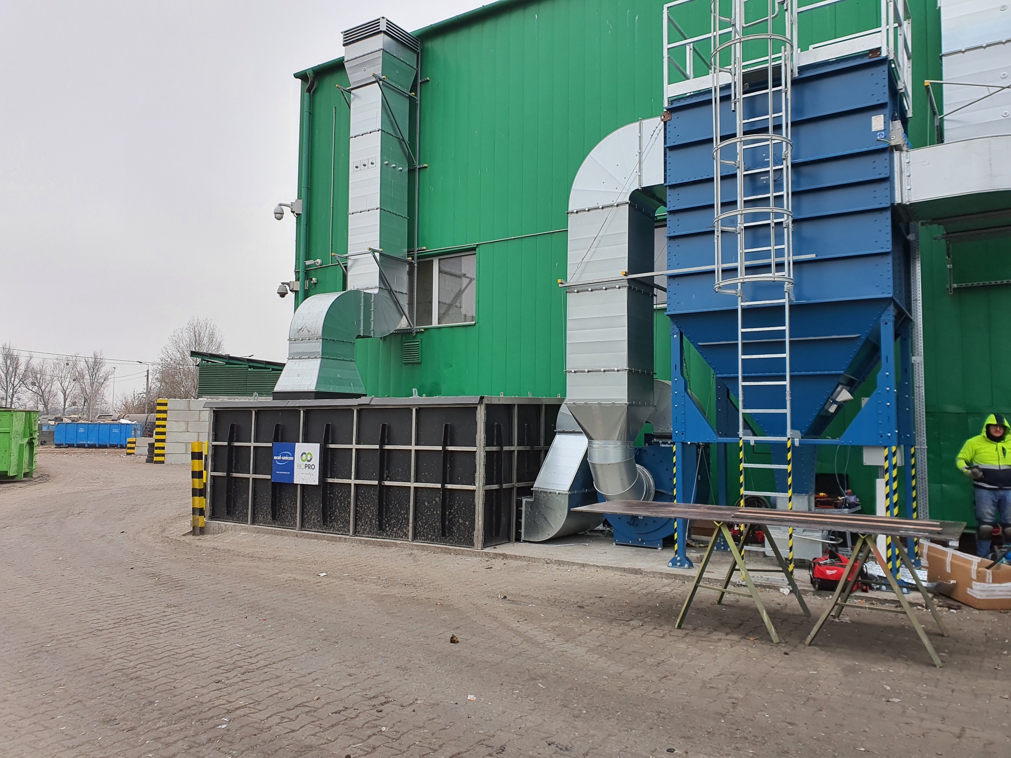 Zewnętrzna instalacja wentylacyjna z rurami i filtrem w kolorze zielonym i niebieskim, ustawiona przy budynku przemysłowym. Widoczny pracownik w odblaskowej kurtce.