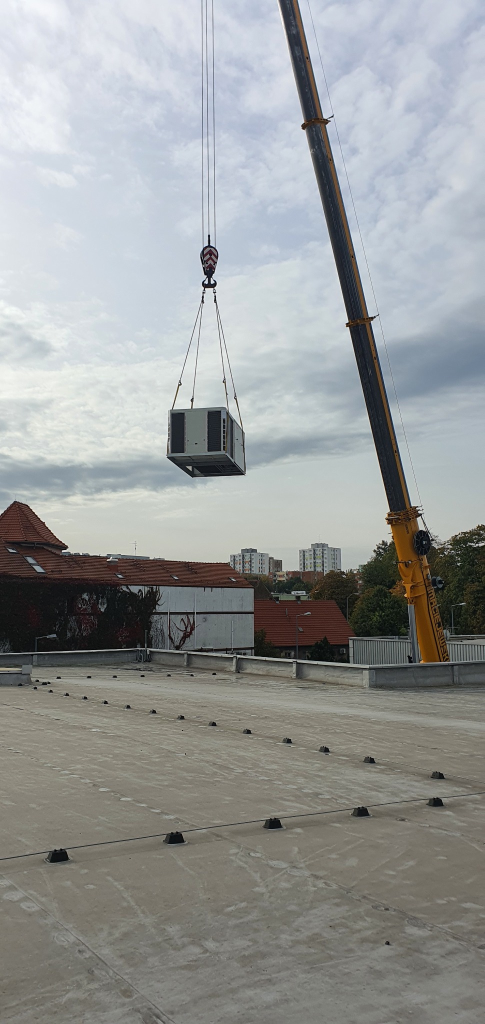 Klimatyzator podnoszony dźwigiem na dach budynku. W tle widać budynki mieszkalne i drzewa. Pochmurne niebo.