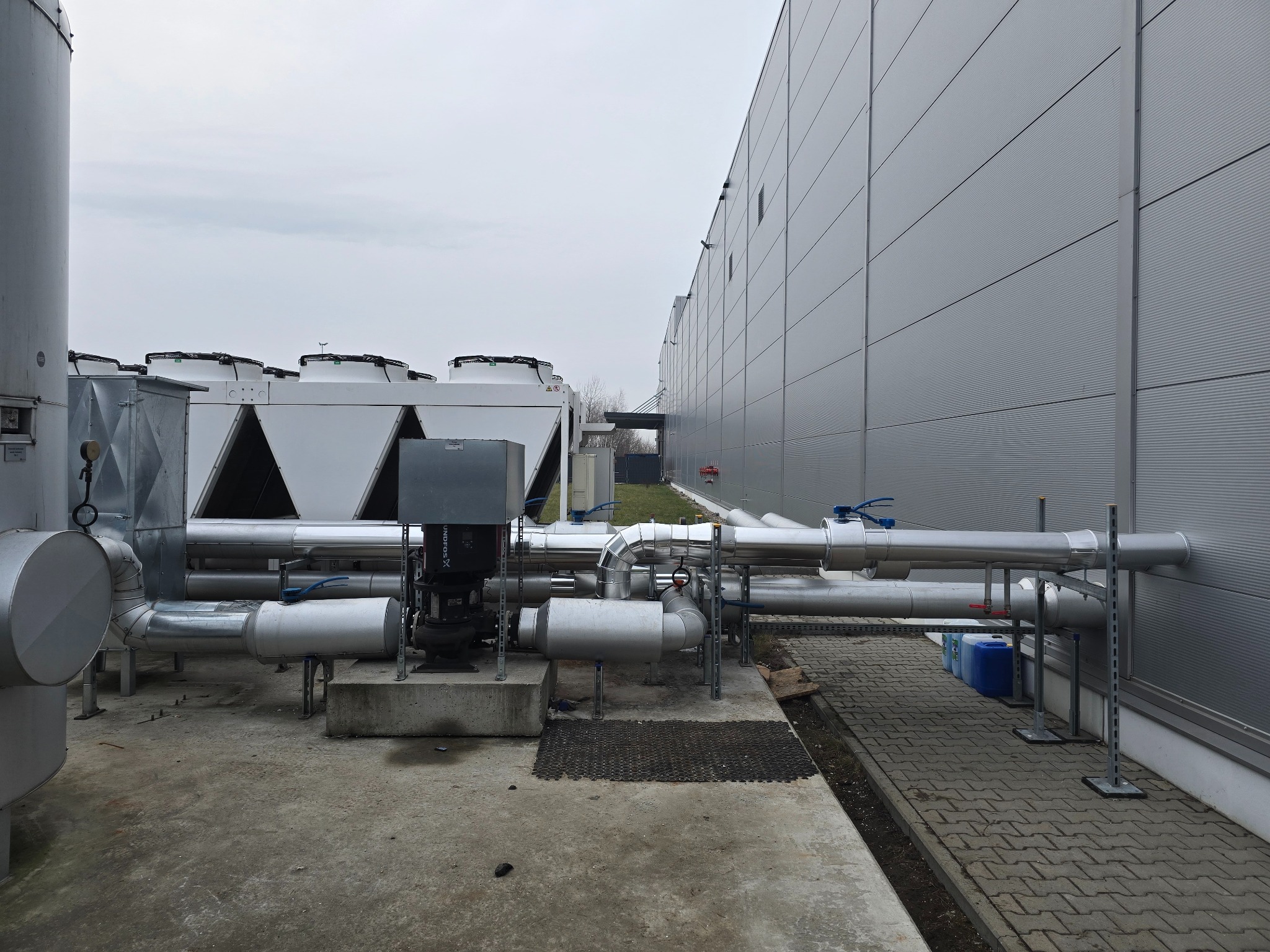 Instalacja przemysłowa z rurami i urządzeniami wentylacyjnymi na dachu budynku. Widoczne elementy chłodnicze i wentylatory. Szara kolorystyka i pochmurne niebo.