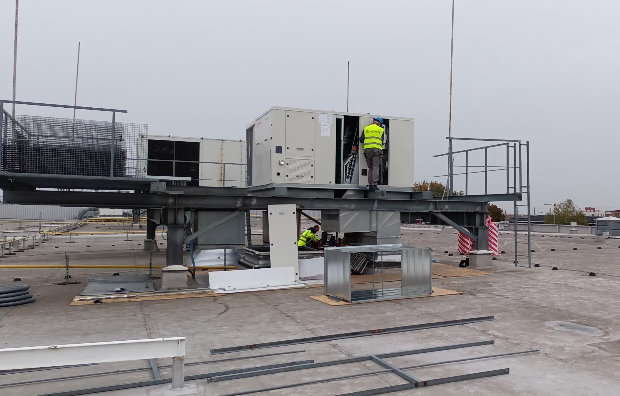 Montaż klimatyzacji przemysłowej na dachu budynku w Łodzi. Dwóch pracowników w odblaskowych kamizelkach instaluje system wentylacyjny na metalowej konstrukcji.