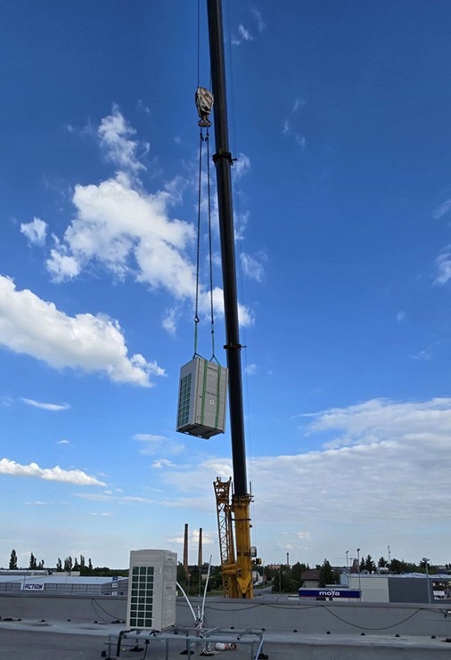 Montaż klimatyzacji na dachu budynku. Żuraw podnosi klimatyzator, widoczny drugi klimatyzator na konstrukcji wsporczej, błękitne niebo z chmurami w tle.