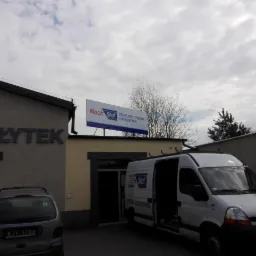 Siedziba firmy Blach Stal, producenta pokryć dachowych, z zaparkowanym białym samochodem dostawczym z otwartymi drzwiami, na tle pochmurnego nieba.