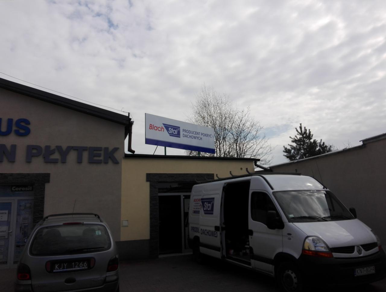 Siedziba firmy Blach Stal, producenta pokryć dachowych, z zaparkowanym białym samochodem dostawczym z otwartymi drzwiami, na tle pochmurnego nieba.