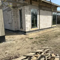 Surowy stan budynku w trakcie budowy z zamontowanymi oknami z czarnymi ramami, widoczne bloczki betonowe, drewniane belki stropowe i stos desek na pierwszym planie.