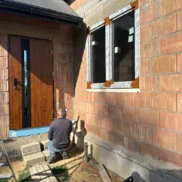 Montaż okna w surowym stanie budynku z cegły, widoczny pracownik na zewnątrz, brązowe okno z białymi ramami ochronnymi, obok drewniane drzwi z czarnym paskiem, kabel zasilający na ziemi.