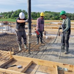 DNB Invest Maciej Klamka - Wylewanie betonu na zbrojenie fundamentów przez trzech robotników w kaskach ochronnych; jeden z nich trzyma wibrator do betonu, tło: budowa w trakcie, zielone drzewa i niebieskie niebo.