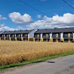 DNB Invest Maciej Klamka - Nowe szeregówki z balkonami w tle pola zboża i błękitnego nieba z chmurami, w oddali widoczny żuraw budowlany.