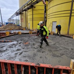 DNB Invest Maciej Klamka - Wylewanie betonu na placu budowy, widoczne szalunki, pracownicy w kaskach i odblaskowych kurtkach, w tle żółte silosy i betoniarka.