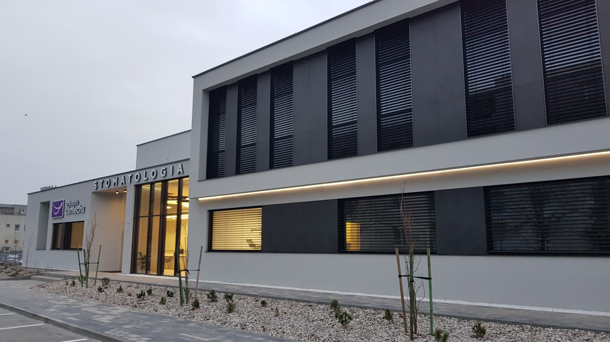 Nowoczesny budynek stomatologii z szarymi panelami elewacyjnymi, żaluzjami zewnętrznymi i podświetlanym napisem 'Stomatologia' nad wejściem.