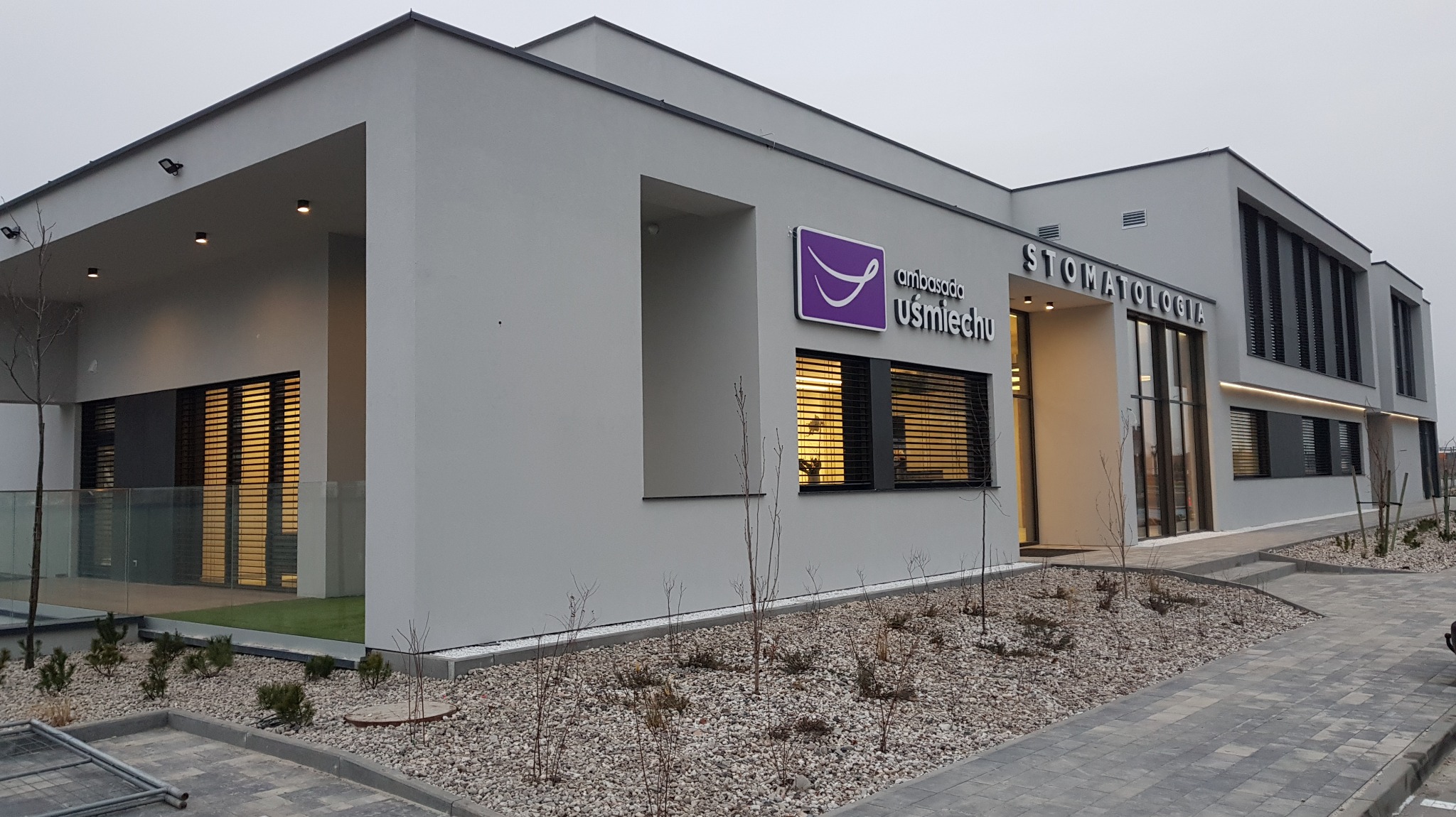 Nowoczesny budynek stomatologii z szarym wykończeniem, logo 'Ambasada Uśmiechu' i zagospodarowanym terenem z kamieniami i młodymi drzewkami.