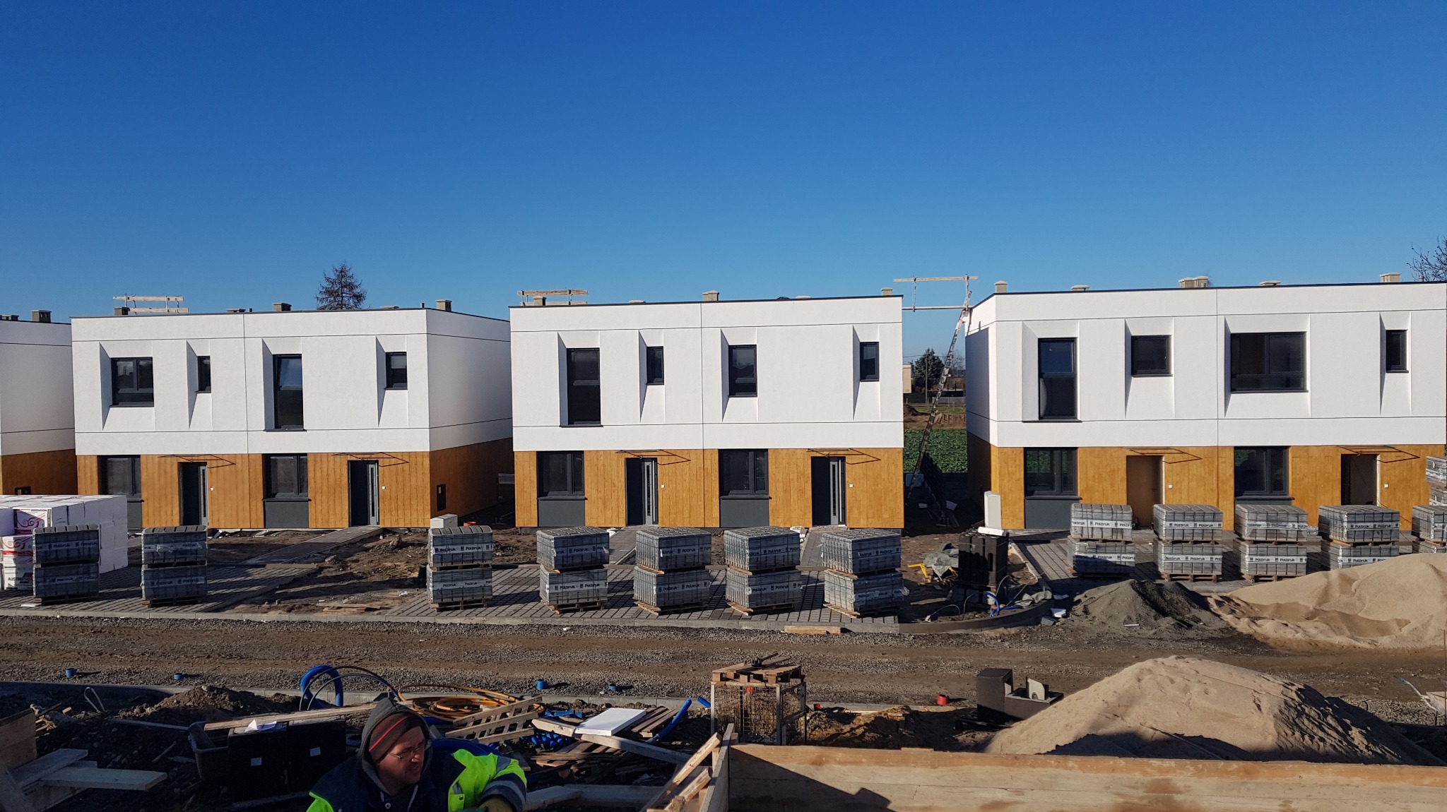 Nowoczesne, szeregowe domy w trakcie budowy z białymi elewacjami i drewnianymi panelami, ułożone kostki brukowe i piasek, widoczny fragment osoby w odblaskowej kamizelce.