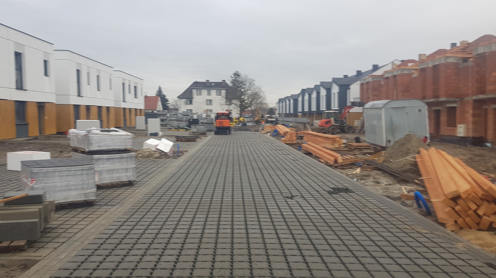 Budowa osiedla domów szeregowych z kostki brukowej, widoczne materiały budowlane i maszyny budowlane w tle, pochmurne niebo.