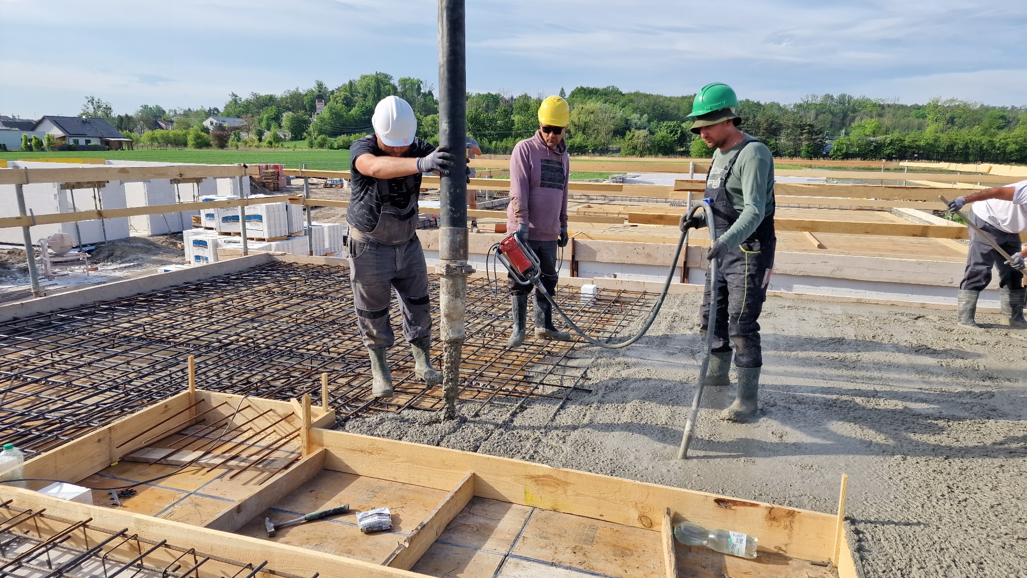 Wylewanie betonu na zbrojenie fundamentów przez trzech robotników w kaskach ochronnych; jeden z nich trzyma wibrator do betonu, tło: budowa w trakcie, zielone drzewa i niebieskie niebo.