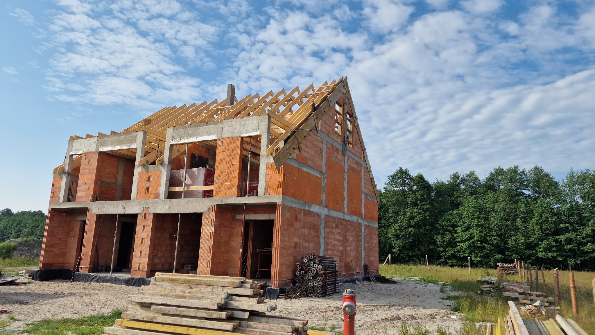 Budowa dwukondygnacyjnego domu z pustaków ceramicznych, widoczne drewniane elementy konstrukcji dachu, w tle las i błękitne niebo z obłokami, na pierwszym planie ułożone deski i czerwony hydrant.