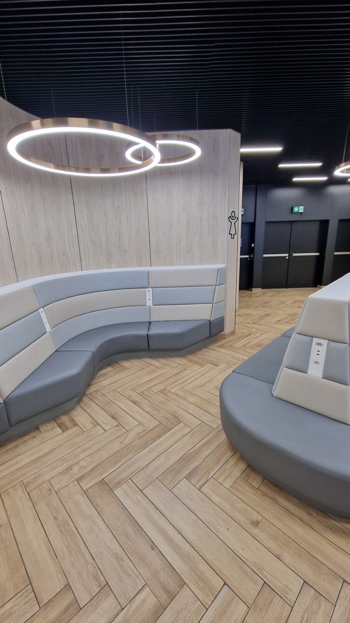Nowoczesna poczekalnia z zakrzywioną sofą w odcieniach szarości i beżu, drewnianą podłogą ułożoną w jodełkę, okrągłymi lampami sufitowymi i oznaczeniem toalety.
