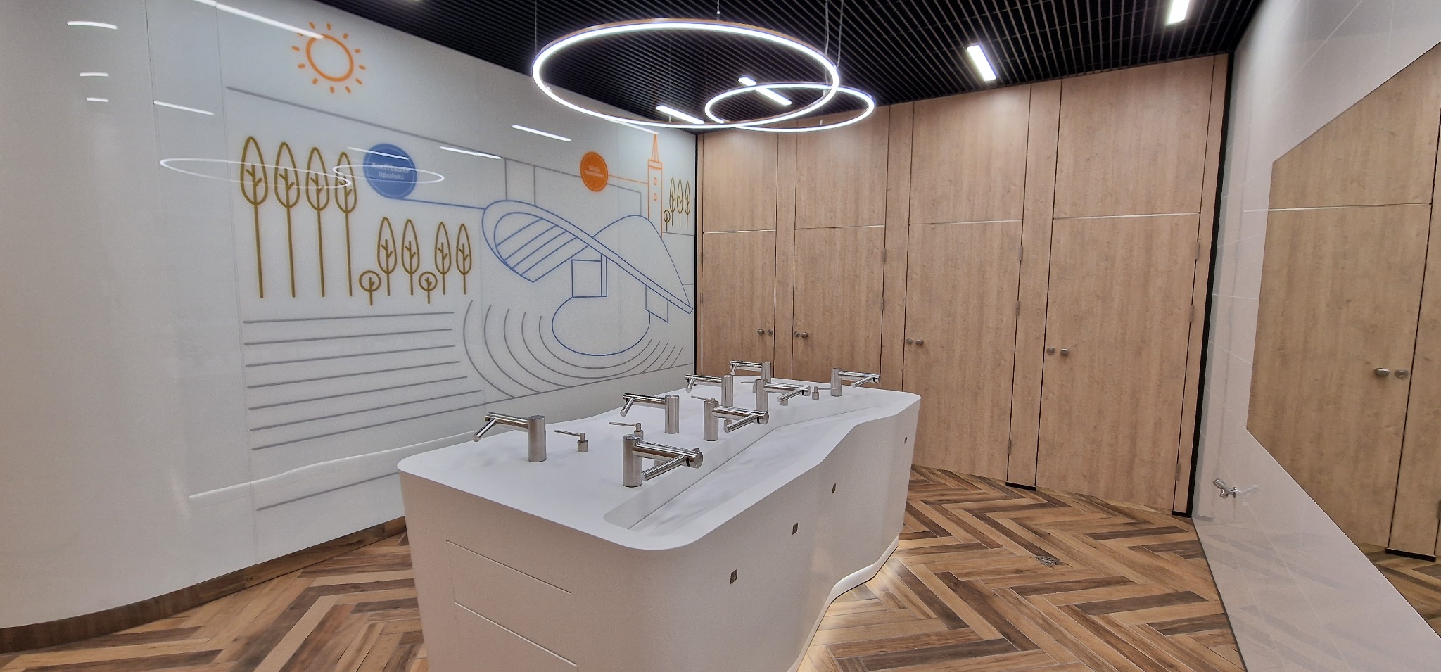 Nowoczesna łazienka publiczna z białym blatem umywalkowym i kilkoma chromowanymi kranami, na ścianie grafika z motywami roślinnymi i architektonicznymi, oświetlenie sufitowe LED w kształcie...