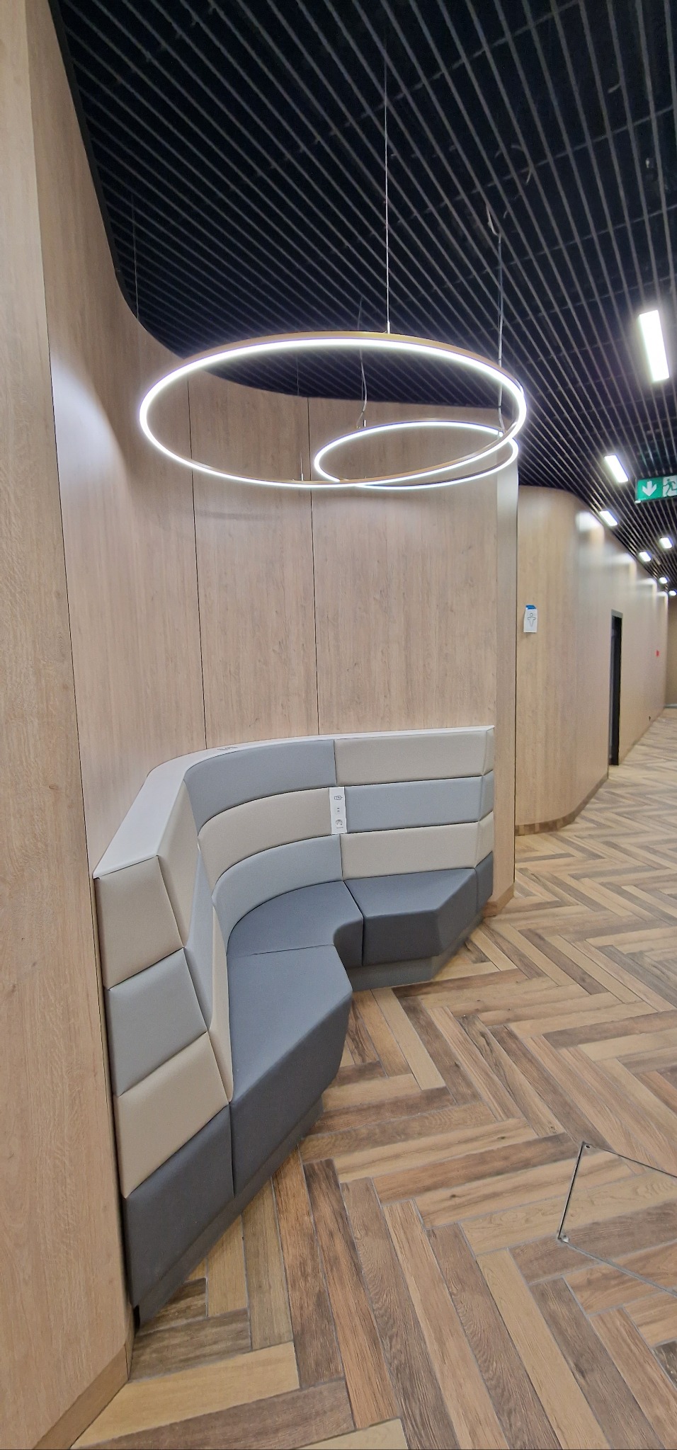 Wnętrze nowoczesnej przestrzeni biurowej z wbudowaną, tapicerowaną ławką w odcieniach szarości i beżu, podświetloną okrągłymi lampami LED zawieszonymi pod czarnym sufitem rastrowym, oraz podłogą...