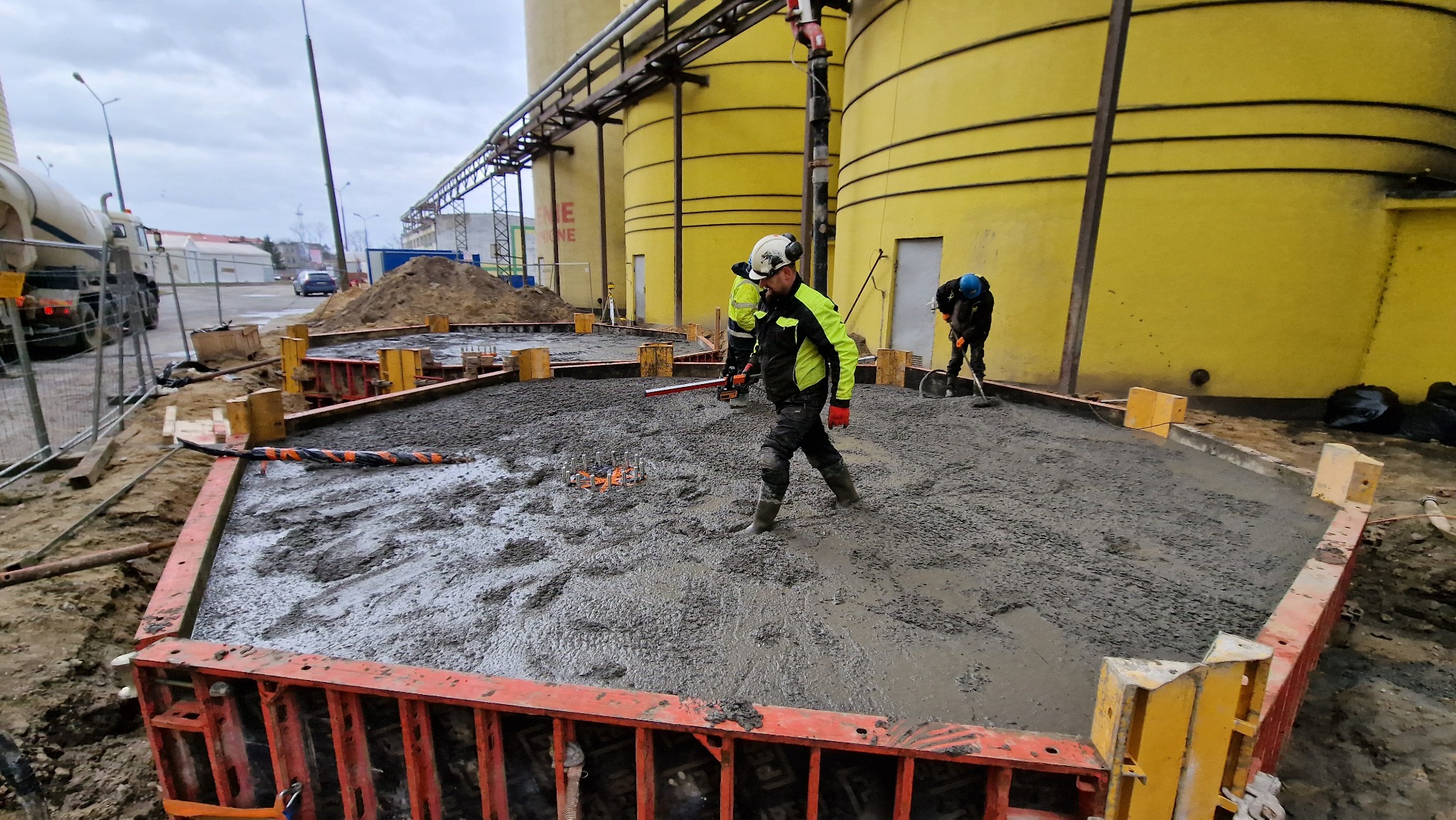 Wylewanie betonu na placu budowy, widoczne szalunki, pracownicy w kaskach i odblaskowych kurtkach, w tle żółte silosy i betoniarka.