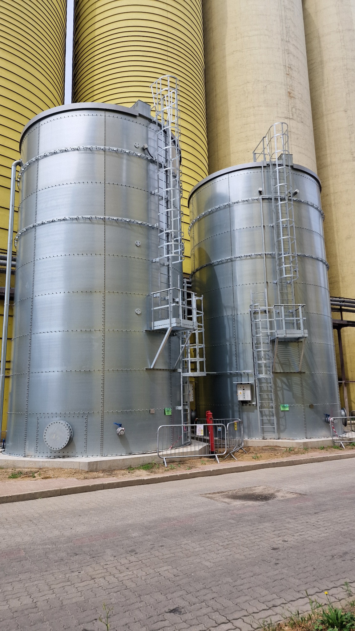 Dwa srebrne, metalowe zbiorniki przemysłowe z drabinkami i platformami, otoczone żółtymi silosami, zabezpieczone barierkami, na tle betonowej nawierzchni.