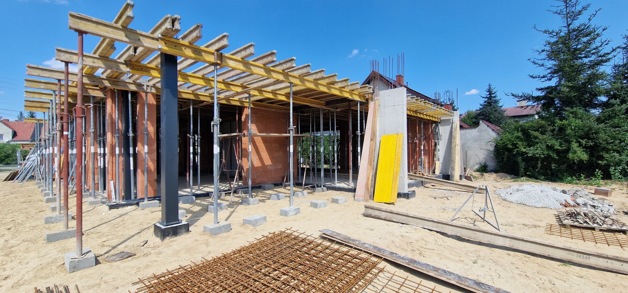 Budowa domu jednorodzinnego w trakcie realizacji: widoczne ściany z pustaków ceramicznych, szalunki stropowe podparte stemplami, zbrojenie fundamentów na piasku oraz stos materiałów budowlanych.