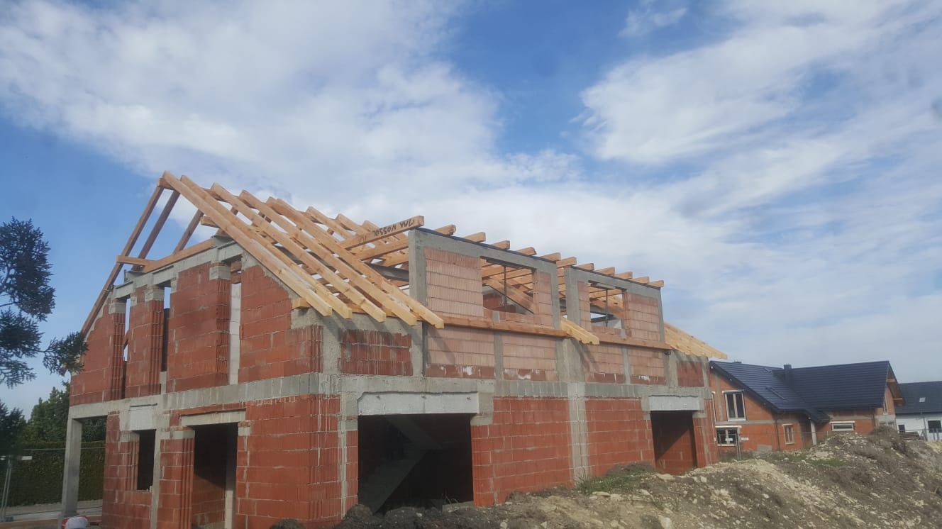 Budowa domu jednorodzinnego w stanie surowym otwartym: cegła, beton, drewniana konstrukcja dachu, widoczne kolejne budynki w tle.