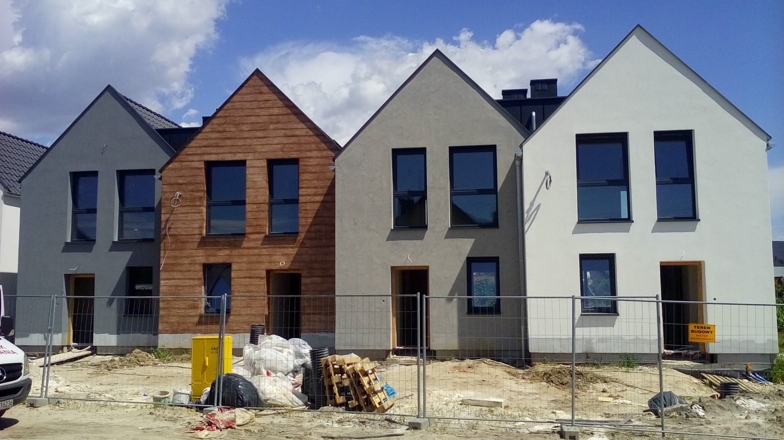 Cztery domy jednorodzinne w różnej fazie wykończenia elewacji: szary, drewniany, szary i biały, zabezpieczone ogrodzeniem tymczasowym, z widocznymi materiałami budowlanymi i żółtą skrzynką gazową.