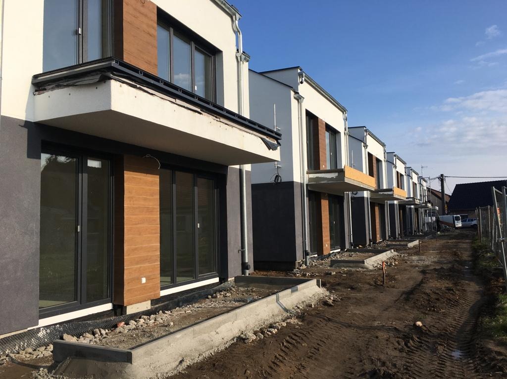 Rząd nowo wybudowanych domów szeregowych z drewnianą elewacją, widoczny stan surowy z przygotowanym terenem pod zagospodarowanie, błękitne niebo.