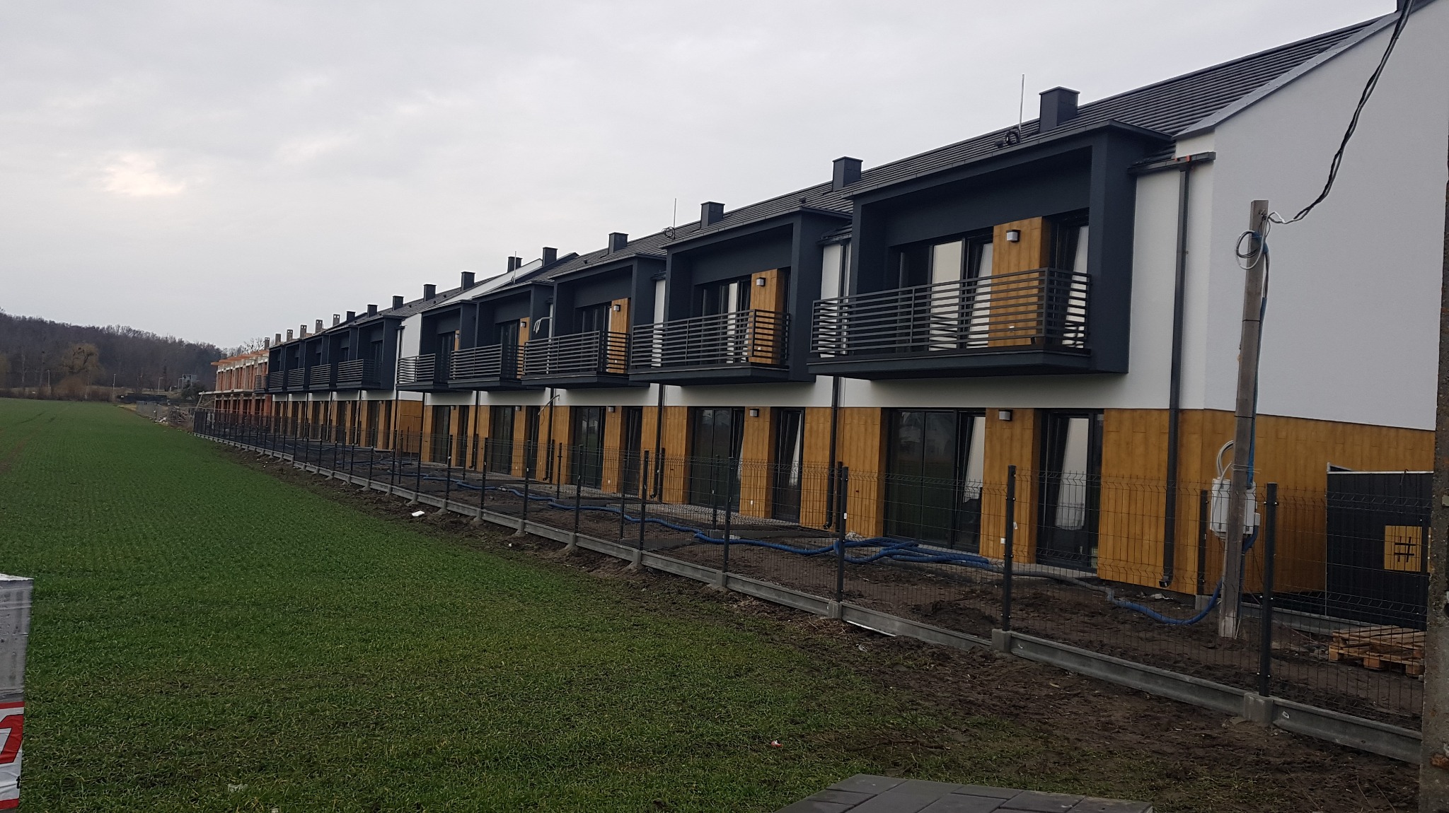 Rząd nowo wybudowanych segmentów mieszkalnych z balkonami, widoczny z perspektywy trawnika przed budynkami, z tymczasowym ogrodzeniem i niebieskimi rurami na ziemi.