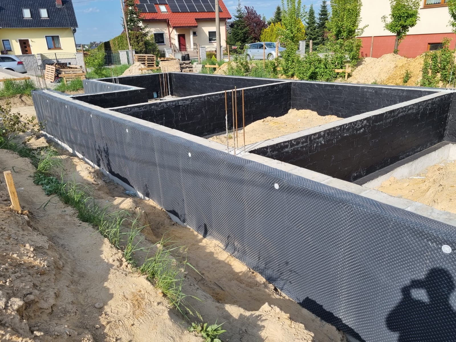 Fundament domu w trakcie budowy z izolacją przeciwwilgociową, widoczne stalowe pręty zbrojeniowe i piasek wewnątrz, w tle budynki mieszkalne