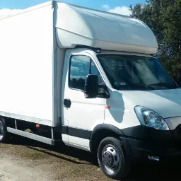 Biały samochód dostawczy Iveco z zabudową kontenerową na tle zieleni, zaparkowany na żwirowej drodze, widoczny z prawej strony. Rejestracja B1 8905U.