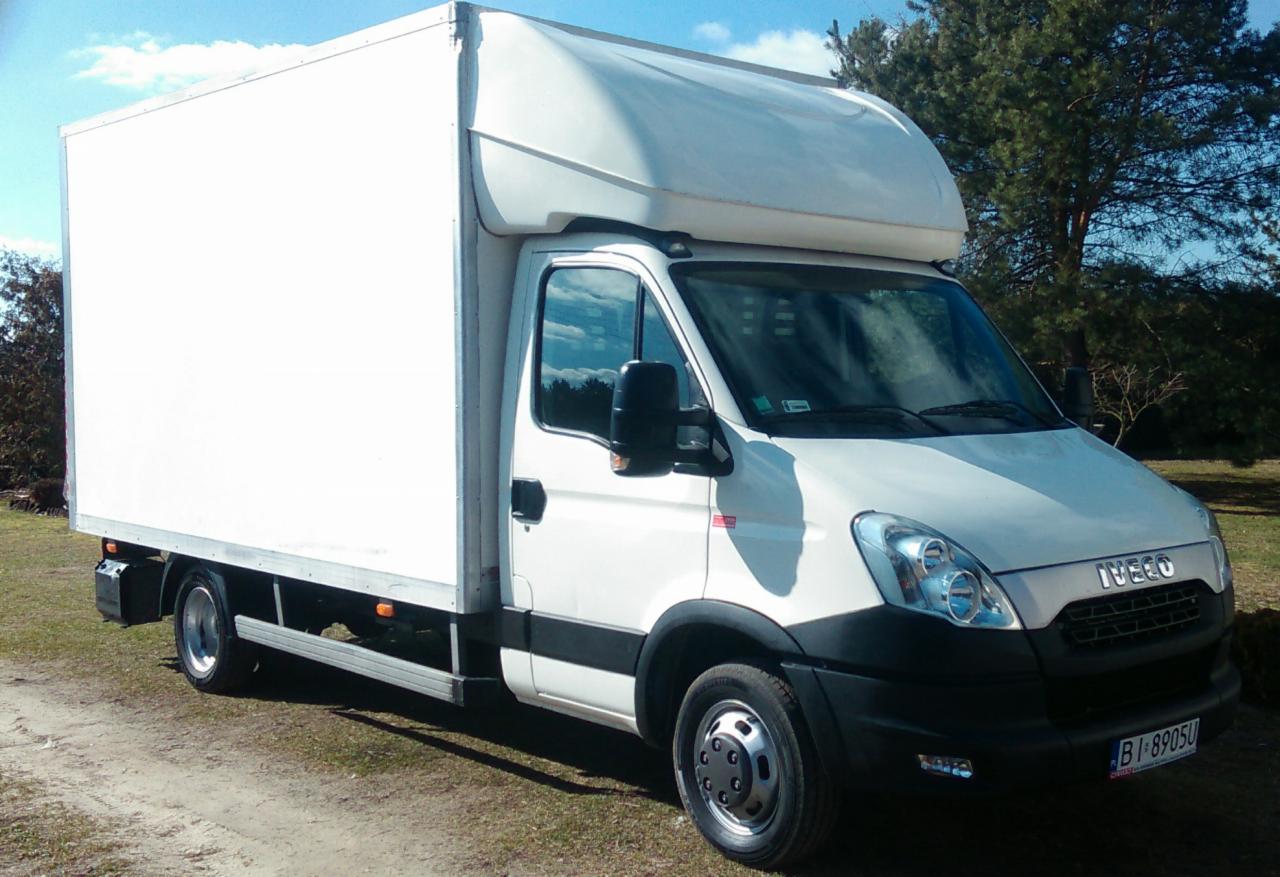 Biały samochód dostawczy Iveco z zabudową kontenerową na tle zieleni, zaparkowany na żwirowej drodze, widoczny z prawej strony. Rejestracja B1 8905U.