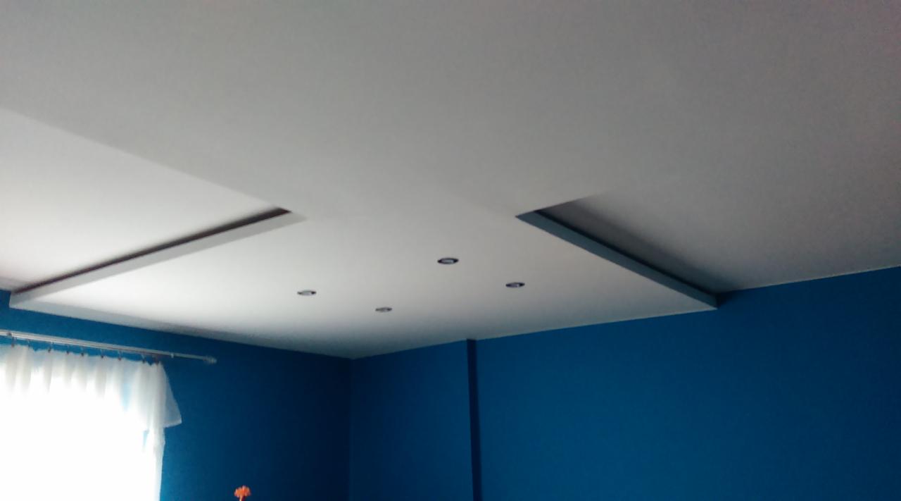 Nowoczesny sufit podwieszany z oświetleniem punktowym w pokoju o ścianach w kolorze głębokiego błękitu. Widok z dołu, fragment okna z firanką po lewej.