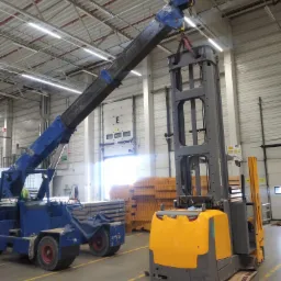 Wózek widłowy Jungheinrich podnoszony przez dźwig w hali magazynowej, widoczne żółte pasy na posadzce i palety w tle.