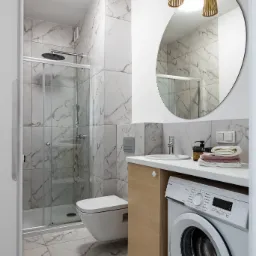 Jasna łazienka z marmurowymi płytkami, kabiną prysznicową, podwieszaną toaletą, umywalką z szafką, pralką Amica i okrągłym lustrem z drewnianą lampą.