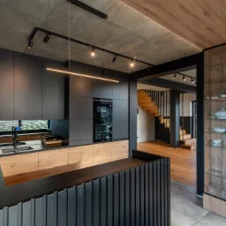 Nowoczesna kuchnia z czarnymi matowymi szafkami, drewnianymi akcentami i betonowym sufitem, oświetlona listwą LED i reflektorami, z widokiem na drewniane schody.