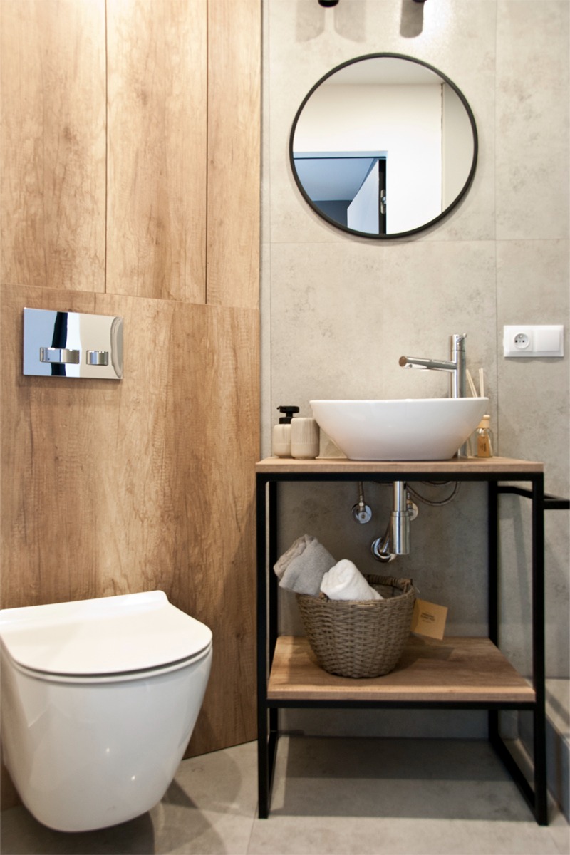 Nowoczesna łazienka z podwieszaną toaletą, umywalką nablatową na metalowym stelażu z drewnianym blatem i okrągłym lustrem w czarnej ramie.