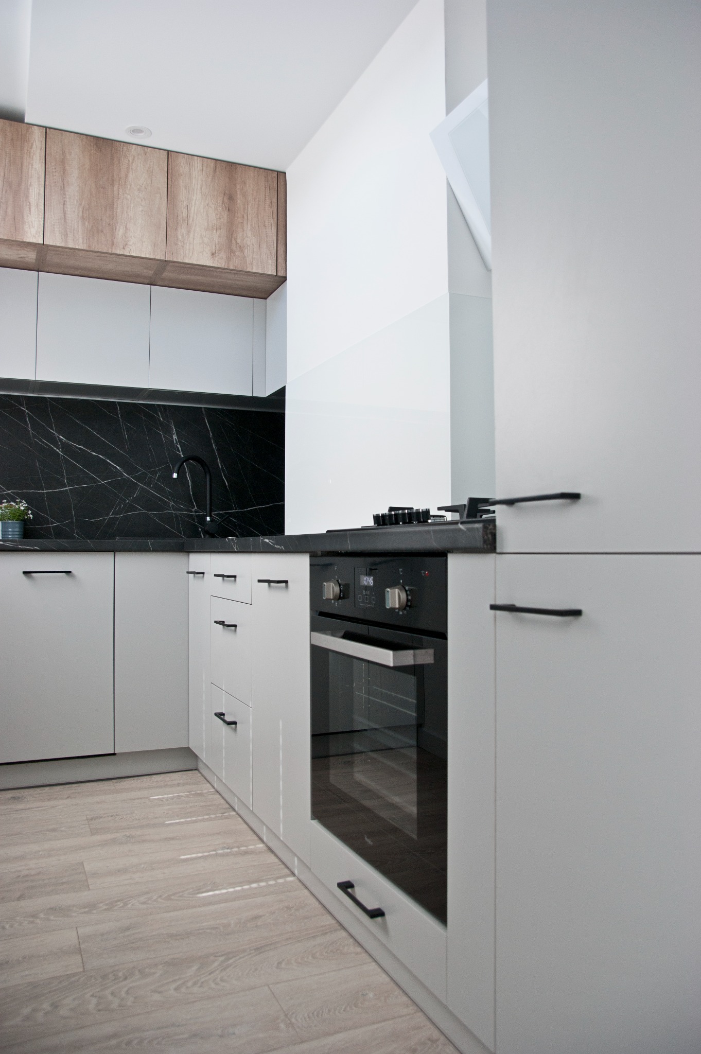 Nowoczesna kuchnia w odcieniach szarości i bieli z czarnym blatem imitującym marmur, drewniane szafki górne, minimalistyczne uchwyty i drewniana podłoga.