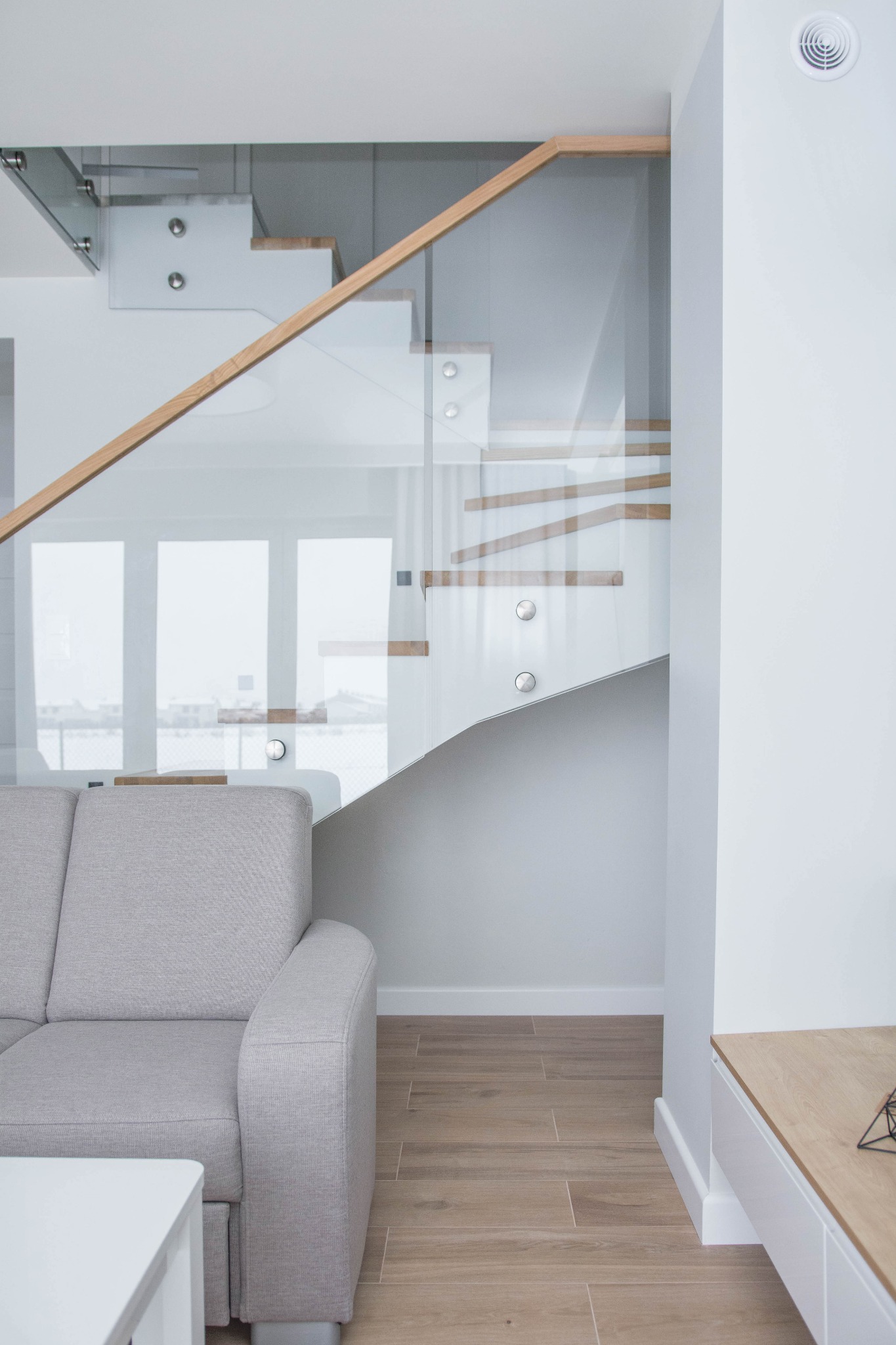 Nowoczesne schody z drewnianymi stopniami i szklaną balustradą w jasnym wnętrzu, widok z perspektywy salonu z szarą sofą i białym stolikiem kawowym.