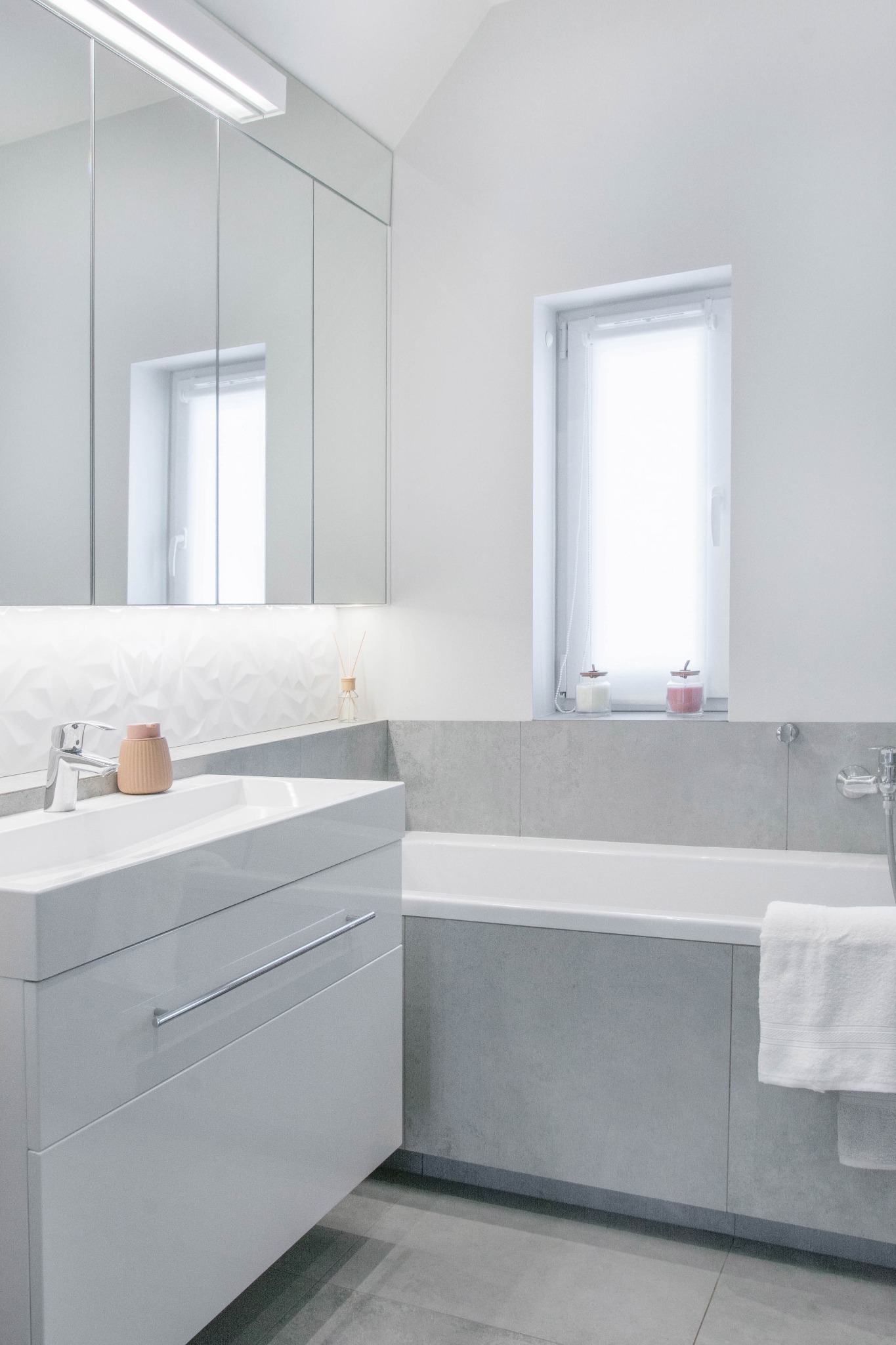 Nowoczesna łazienka w odcieniach szarości i bieli, z minimalistyczną umywalką z szafką, wanną obudowaną płytkami i lustrzaną szafką nad umywalką, uzupełniona o dekoracyjne akcenty i oświetlenie LED.